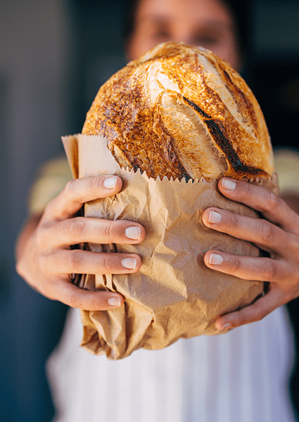 Hands offering crusty sourdough loaf in bag
